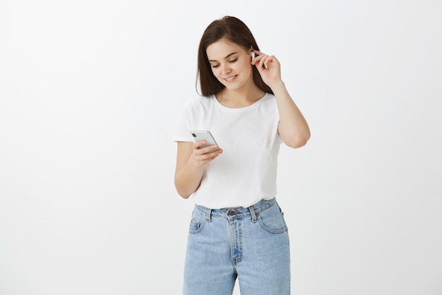 Jeune femme posant avec son téléphone et écouteurs contre le mur blanc