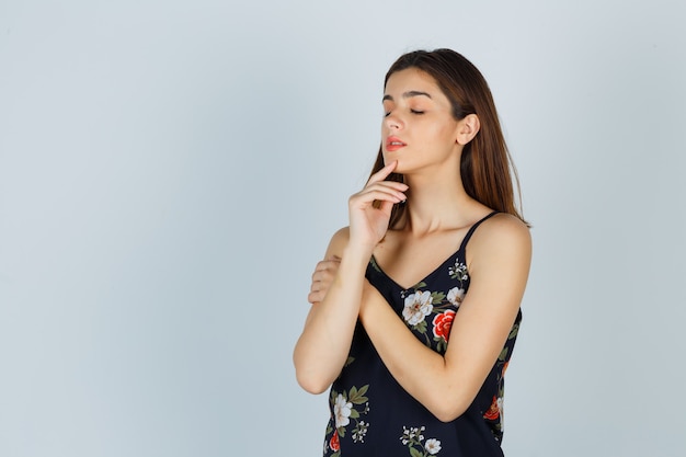 Jeune femme posant tout en touchant la peau du visage sur son menton en blouse et l'air délicieuse, vue de face.