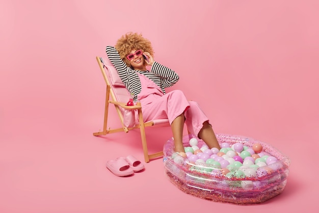 Jeune femme positive aux cheveux bouclés pose sur une chaise longue confortable fait un appel téléphonique porte des lunettes de soleil pull rayé et une salopette rêve de vacances d'été isolées sur fond rose