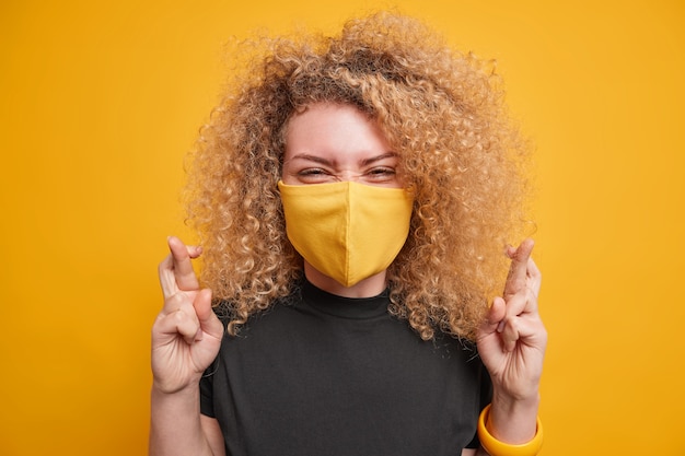 Une jeune femme positive a les cheveux bouclés et croise les doigts anticipe de bons résultats positifs espère que les rêves deviennent réalité porte un t-shirt noir et un masque jetable pour empêcher la propagation du virus isolé sur jaune