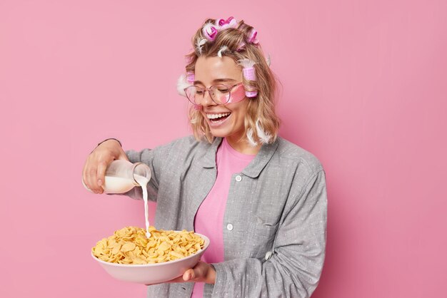 Une jeune femme positive fait une coiffure avec des rouleaux qui vont prendre le petit déjeuner verse du lait dans des cornflakes vêtus de vêtements de nuit subit des procédures de beauté isolées sur fond rose Heure du matin