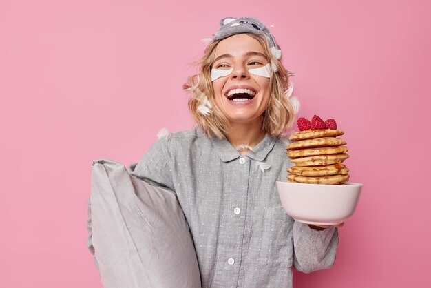 Une jeune femme positive et insouciante s'amuse à se réveiller le matin en riant joyeusement vêtue de vêtements de nuit applique des patchs de beauté sous les yeux tient un oreiller moelleux et une pile de crêpes a des plumes coincées dans les cheveux