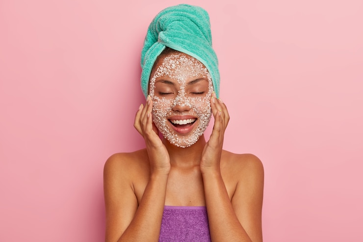 Photo gratuit une jeune femme positive masse le visage avec un gommage spécial, réduit les points noirs sur les joues, ressent le plaisir des soins de beauté, a des problèmes de peau, se soucie des cheveux, enveloppée dans une serviette. haute résolution