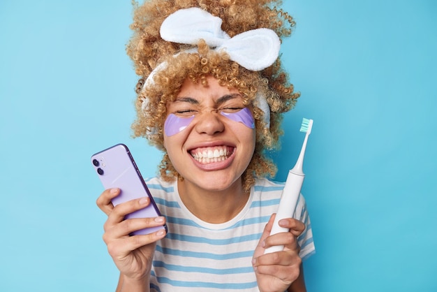 Une jeune femme positive montre des dents blanches après le nettoyage prend soin de la peau applique des patchs de beauté contient une brosse à dents électrique moderne et un smartphone porte un t-shirt à rayures bandeau isolé sur un mur bleu