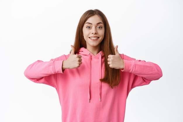 Une jeune femme positive montre son pouce levé, approuve et aime un bon produit, accepte ou paise le choix, se dresse sur fond blanc