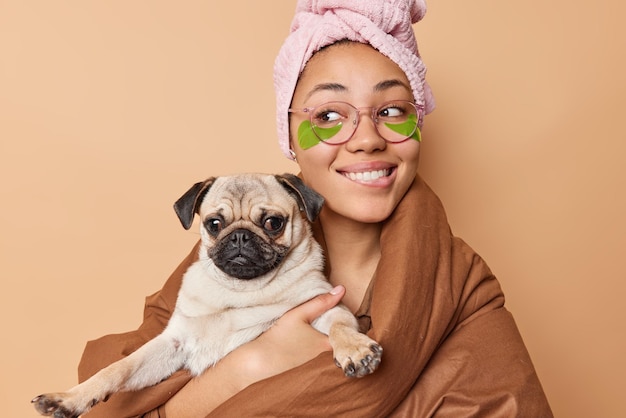 Une jeune femme positive mord les lèvres regarde joyeusement loin des poses avec un chien carlin enveloppé dans une couverture porte une serviette sur la tête applique des patchs de beauté verts pour réduire les rides isolées sur fond marron