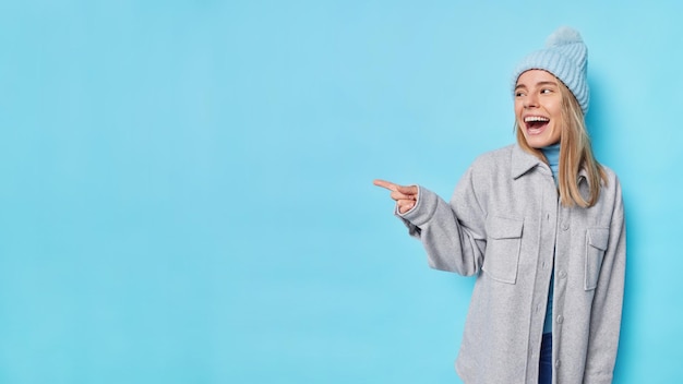 Photo gratuite une jeune femme positive et optimiste rit joyeusement en pointant l'index vers l'extérieur montre une belle publicité porte un chapeau et une veste grise isolée sur fond bleu regardez cette offre ou cette réduction incroyable