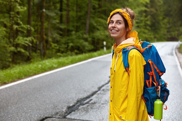 Jeune femme positive avec sac à dos, se tient sur le côté à la caméra, traverse la route