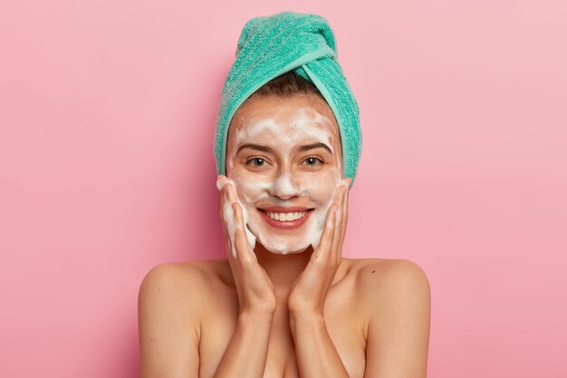 Une jeune femme positive a le sourire à pleines dents, a des dents parfaites, tapote la peau avec du savon sanitaire liquide, se lave avec un gel moussant, se réveille le matin pour avoir une routine de beauté