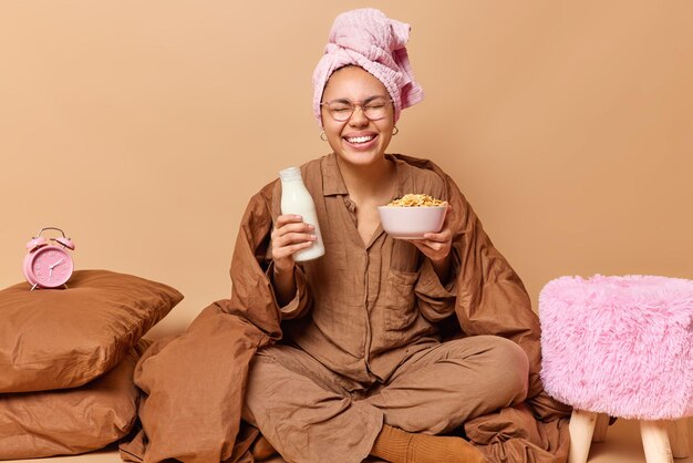 Une jeune femme positive vêtue d'un costume de sommeil tient une bouteille de lait frais et un bol de céréales sourit joyeusement recouvert de poses de couverture sur le lit prend un petit déjeuner sain isolé sur fond beige