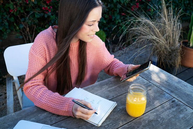 Photo gratuit jeune femme prenant des notes à l'extérieur tout en utilisant un smartphone
