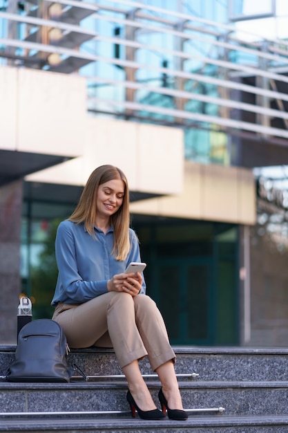 Jeune femme professionnelle assise sur l'escalier en face du bâtiment en verre, parler au téléphone mobile