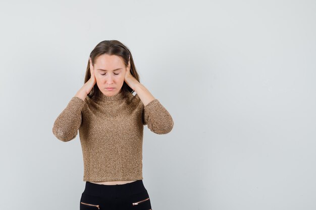 Jeune femme en pull doré or et pantalon noir en appuyant sur les mains sur les oreilles et à la vue épuisée, de face.