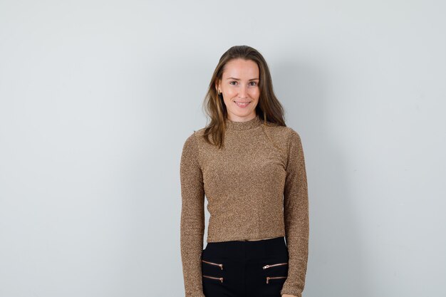 Jeune femme en pull doré et pantalon noir debout droit et souriant et à la recherche de plaisir