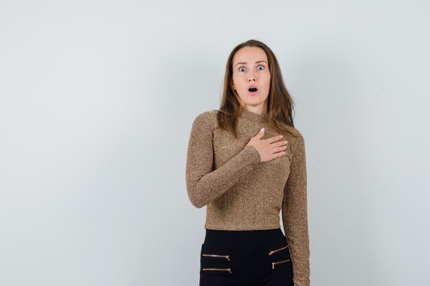 Jeune femme en pull doré et pantalon noir tenant la main sur la poitrine et à la surprise