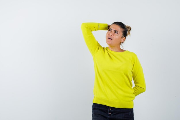 Jeune femme en pull jaune et pantalon noir mettant la main sur la tête tout en tenant la main derrière la taille et en regardant pensive