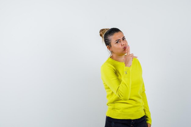 Jeune femme en pull jaune et pantalon noir montrant le geste du pistolet et l'air confiant