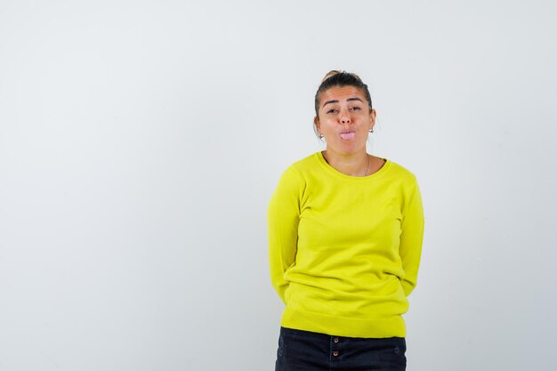 Jeune femme en pull jaune et pantalon noir se tenant droit, grimaçant et posant devant la caméra et ayant l'air heureux