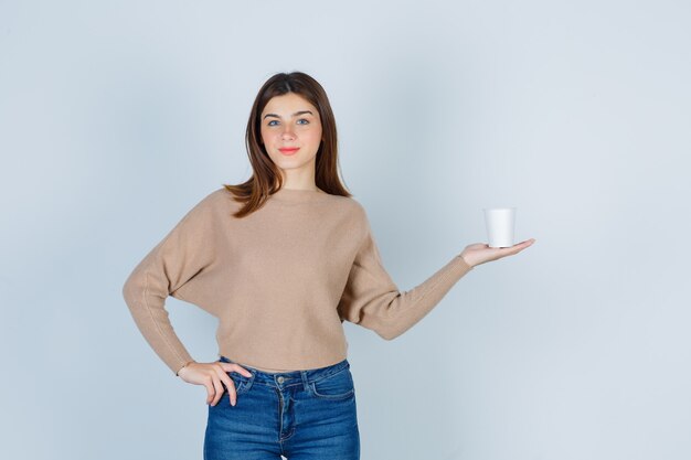 Jeune femme en pull, jeans avec la main sur la taille, gardant une tasse de papier et l'air heureux, vue de face.