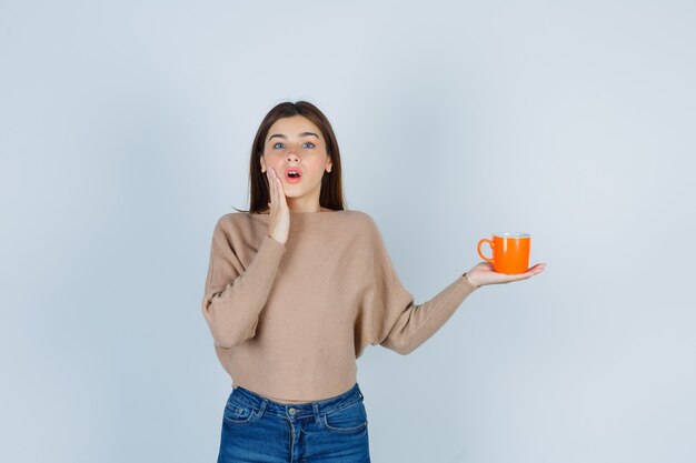 Jeune femme en pull, jeans montrant la coupe, avec la main sur la joue et l'air choqué, vue de face.