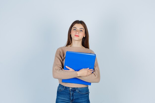 Jeune femme en pull, jeans regardant à l'avant, tenant un dossier et l'air pensif, vue de face.