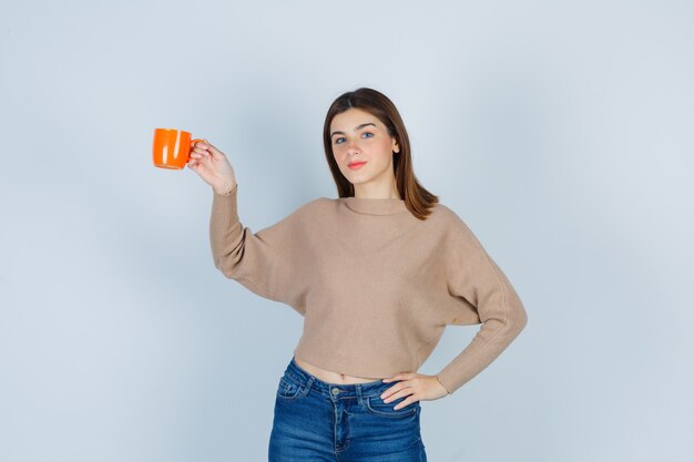 Jeune femme en pull, jeans soulevant une tasse, avec la main sur la taille et l'air réfléchi, vue de face.