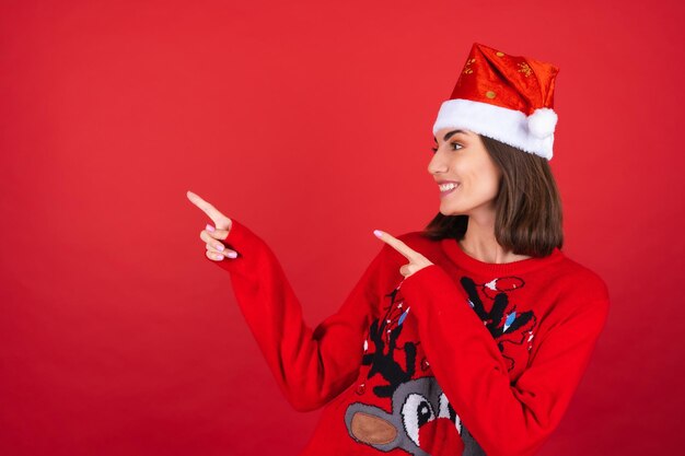 Jeune femme en pull de noël et bonnet de noel souriant avec enthousiasme, pointant son doigt vers la gauche vers un espace vide