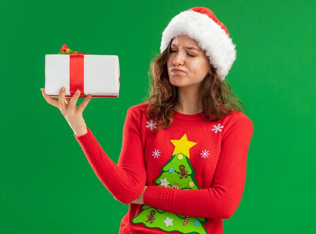 Jeune femme en pull de Noël rouge et bonnet de Noel tenant un cadeau en le regardant avec une expression sceptique debout sur fond vert