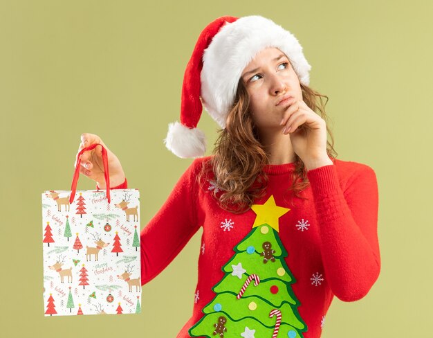 Jeune femme en pull de noël rouge et bonnet de noel tenant un sac en papier avec des cadeaux de noël levant la pensée avec la main sur son menton debout sur un mur vert