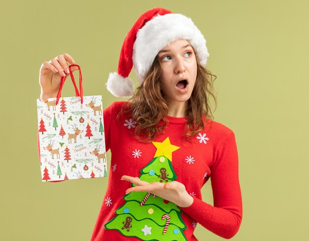 Jeune femme en pull de noël rouge et bonnet de noel tenant un sac en papier avec des cadeaux de noël se présentant avec le bras l'air surpris debout sur un mur vert