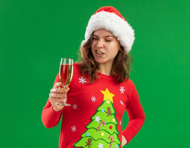 Jeune femme en pull de Noël rouge et bonnet de noel tenant un verre de champagne en le regardant confus debout sur fond vert