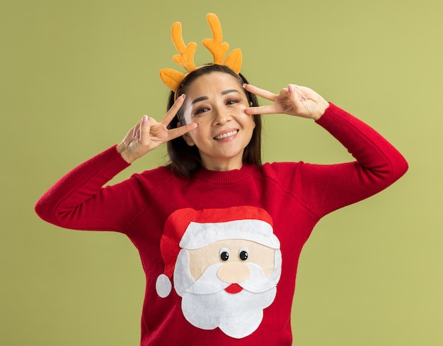 Jeune Femme En Pull De Noël Rouge Portant Une Jante Drôle Avec Des Cornes De Cerf Regardant La Caméra En Souriant Avec Un Visage Heureux Montrant Un Signe V Près De Ses Yeux Debout Sur Fond Vert