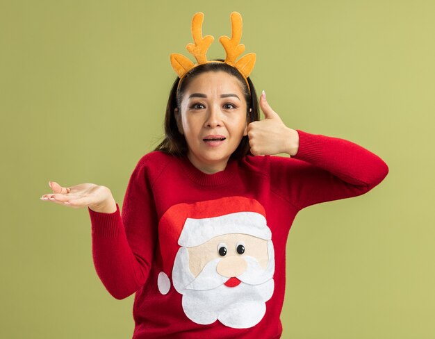 Jeune femme en pull de Noël rouge portant une jante drôle avec des cornes de cerf à la surprise montrant les pouces vers le haut présentant l'espace de copie avec le bras de la main