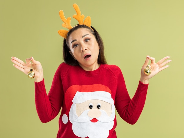 Jeune femme en pull de noël rouge portant une jante drôle avec des cornes de cerf tenant des boules de noël confuses n'ayant pas de réponse debout sur un mur vert