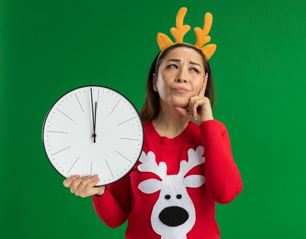 Jeune femme en pull de Noël rouge portant une jante drôle avec des cornes de cerf tenant horloge murale à la perplexité debout sur fond vert
