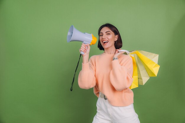 Jeune femme en pull pêche décontracté isolé sur un mur de couleur vert olive cri dans un mégaphone tenant des sacs à provisions, annonce des remises promotion de vente