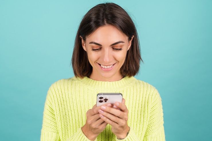Photo gratuit jeune femme en pull vert clair avec un téléphone portable avec un message de saisie de sourire