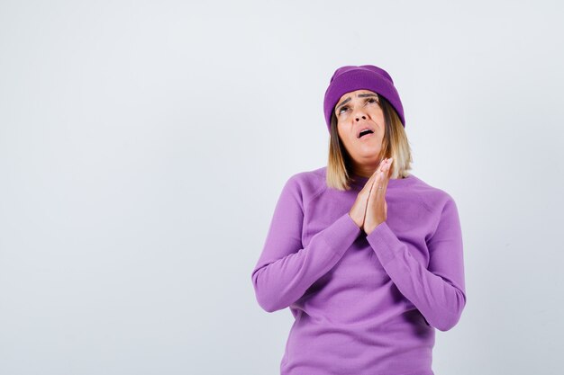 Jeune Femme En Pull Violet, Bonnet Avec Les Mains En Geste De Prière Et L'air Plein D'espoir, Vue De Face.