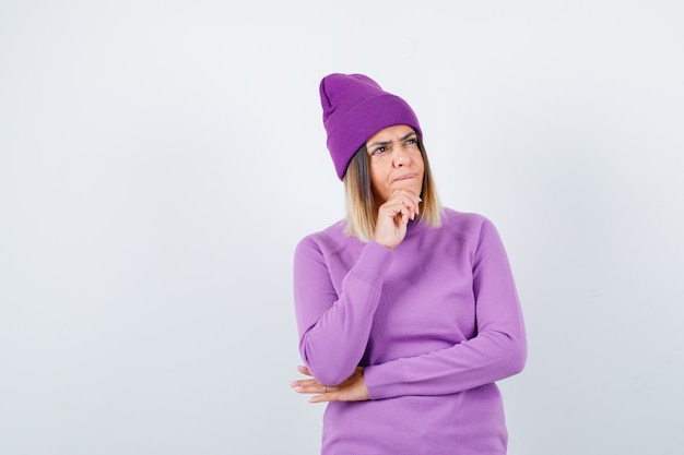 Jeune femme en pull violet, bonnet soutenant le menton à portée de main et pensif, vue de face.