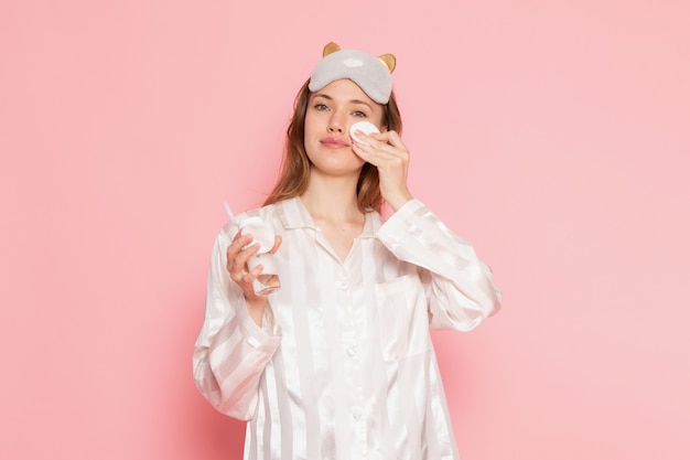 Jeune femme en pyjama et masque de sommeil nettoyant son visage sur rose