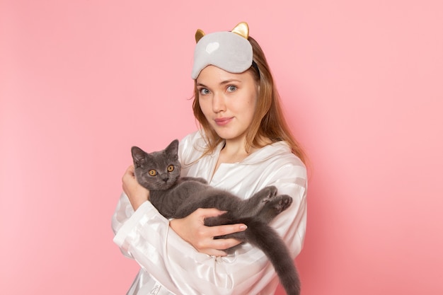 jeune femme en pyjama et masque de sommeil posant avec un léger sourire et chat sur rose