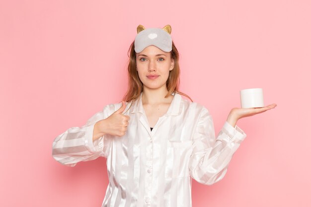 jeune femme en pyjama et masque de sommeil souriant et tenant la crème sur rose