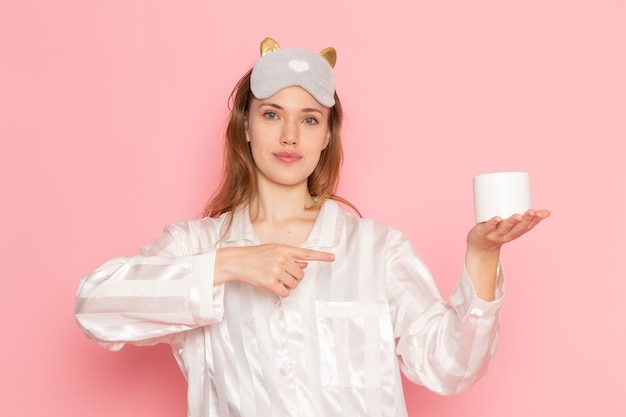 Jeune femme en pyjama et masque de sommeil tenant la crème blanche peut sur rose