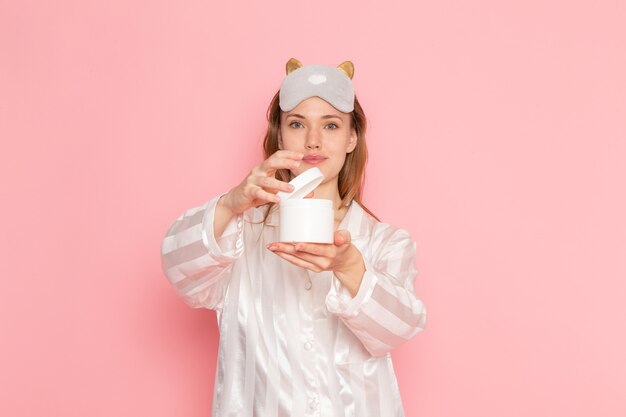 Jeune femme en pyjama et masque de sommeil tenant peut de crème sur rose