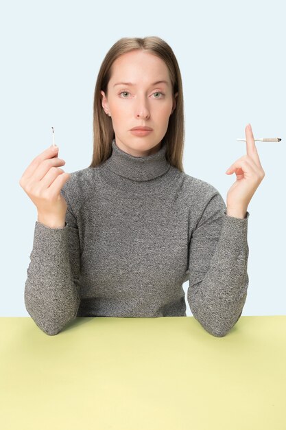 La jeune femme qui fume la cigarette alors qu'elle était assise à table au studio. Couleurs à la mode. Le portrait de fille caucasienne dans un style minimalisme avec espace copie