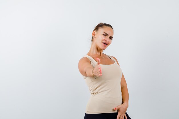 Jeune femme qui s'étend de la main pour saluer en débardeur beige et à la confiance. vue de face.