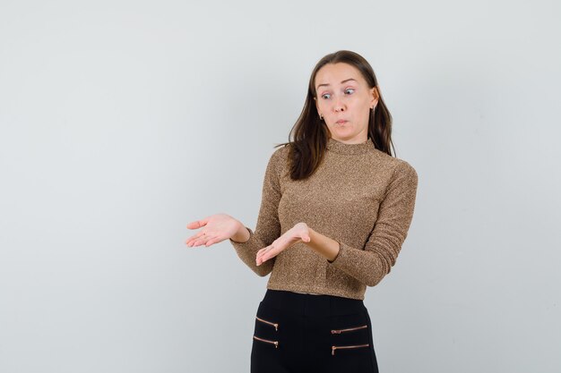 Jeune femme qui s'étend les mains afin de recevoir quelque chose en pull doré et pantalon noir et à la surprise, vue de face.