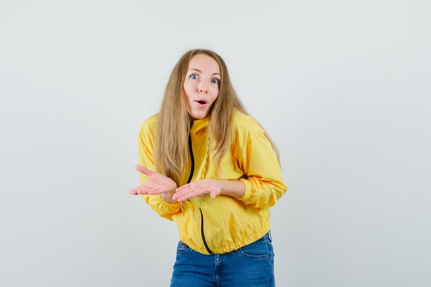 Jeune femme qui s'étend les mains comme expliquant quelque chose en blouson aviateur jaune et jean bleu et à l'optimiste, vue de face.