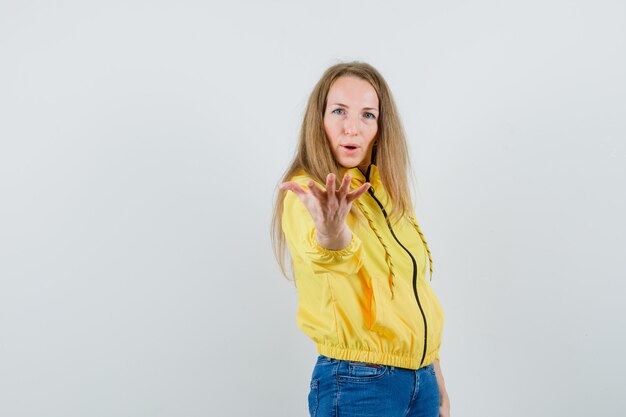 Jeune femme qui s'étend les mains comme invitant à venir en blouson aviateur jaune et jean bleu et à l'optimiste, vue de face.