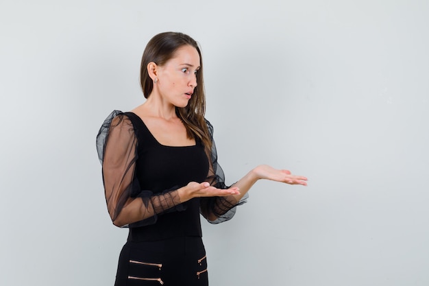 Jeune femme qui s'étend les mains pour recevoir quelque chose en chemisier noir et pantalon noir et à la vue de face, choquée.